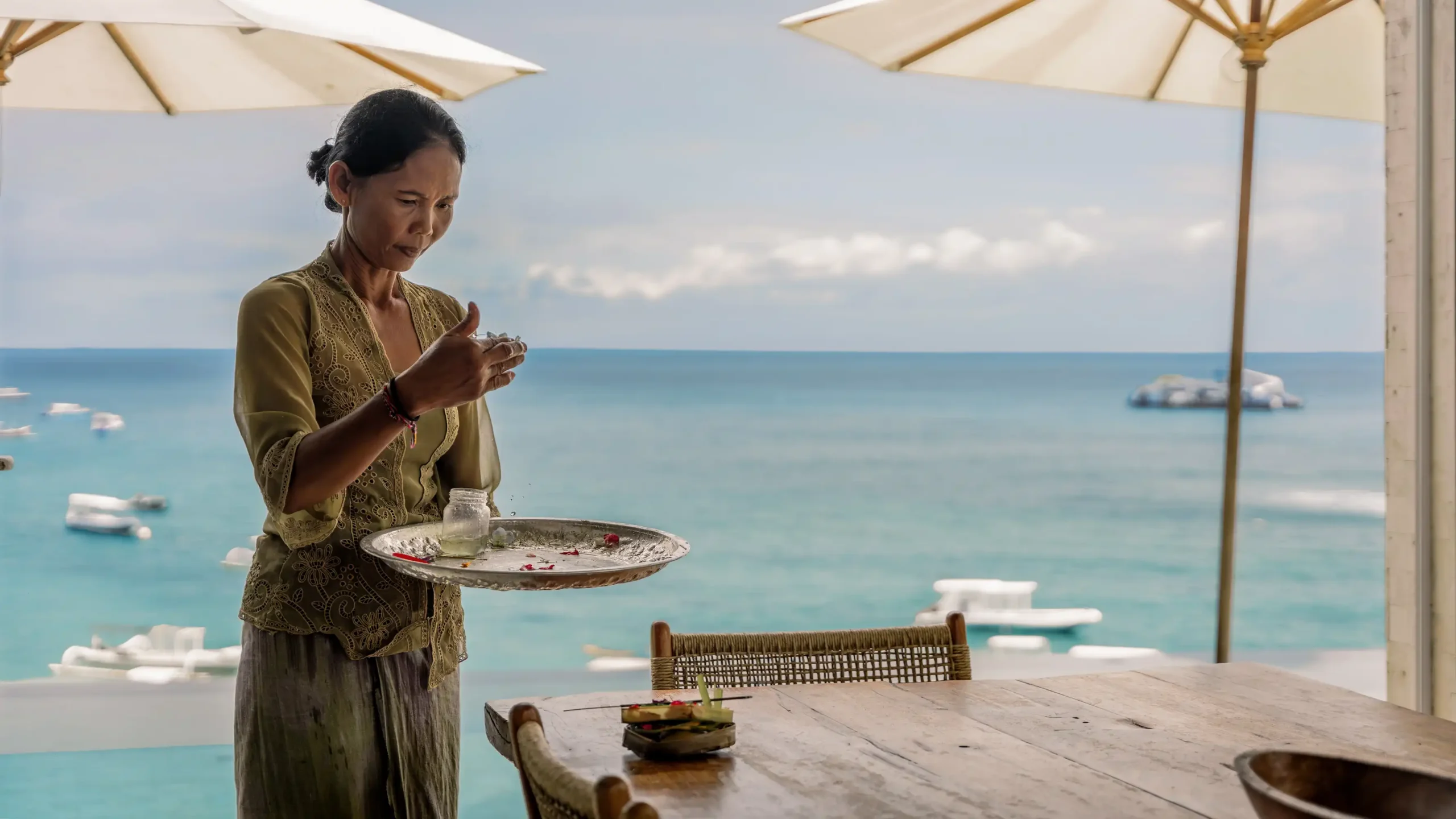 "Beach House staff plating breakfast on the outdoor dining table with jukungs offshore at Lago Nusa Lembongan