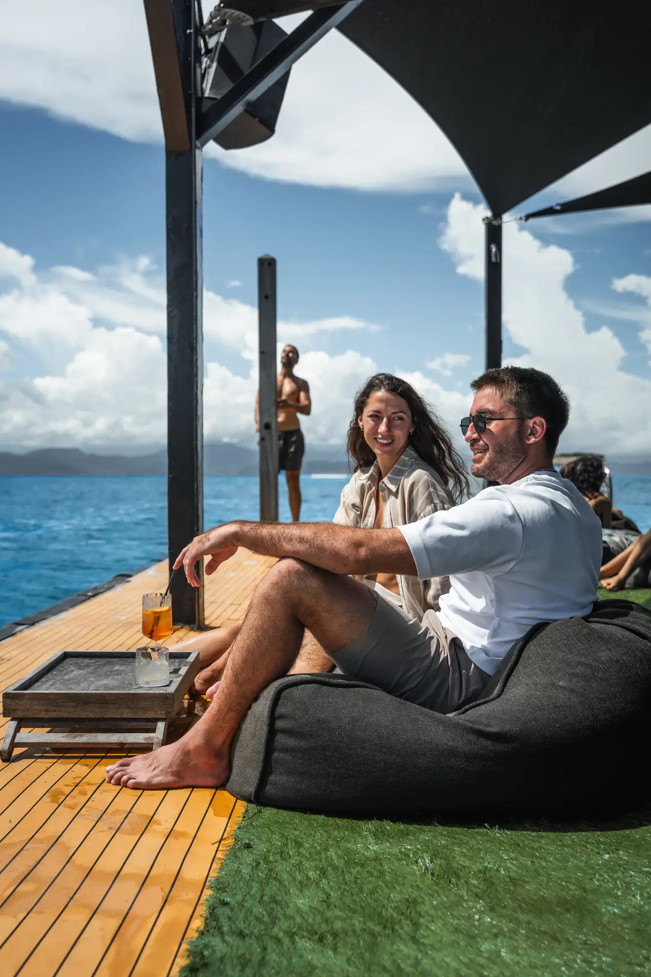 A couple lounges on a charcoal bean bag on the Pontoon at Lago deck, an Aperol spritz on the side table and the open ocean stretching behind.