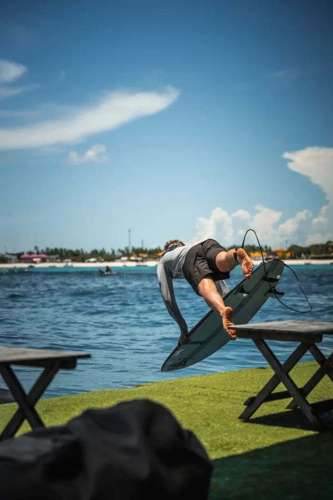 A surfer leaps from the astroturf deck of the Pontoon at Lago with board in hand, crystal-clear water below and the palm-fringed Lembongan shore beyond.